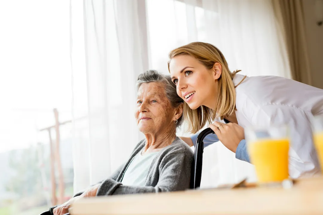 A caregiver leaning beside an older woman in a wheelchair by a sunny window